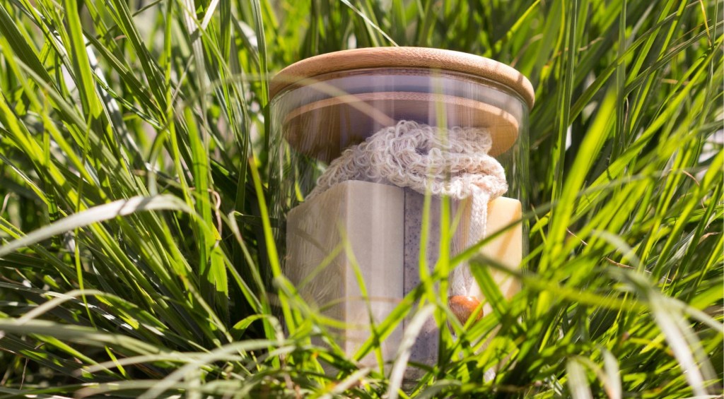 Bocal en verre avec couvercle en bois contenant des savons artisanaux et une pochette en sisal, posé dans l'herbe haute.