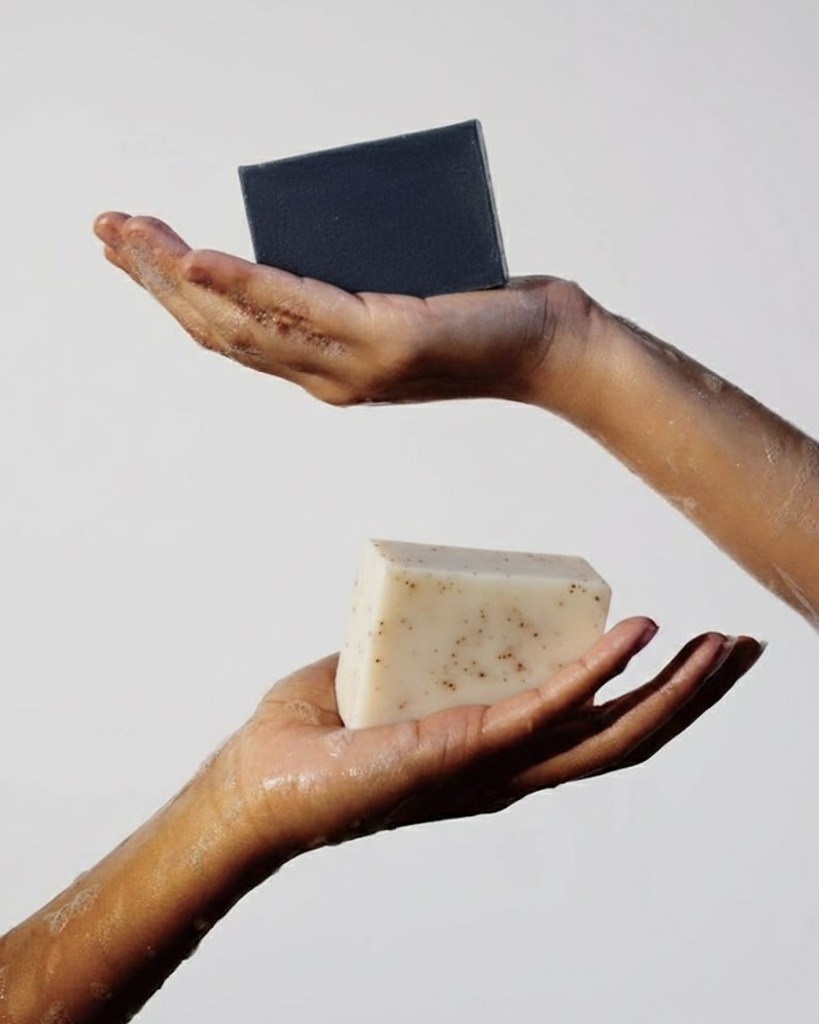 Deux mains tenant chacune un savon : un savon noir au charbon et un savon blanc exfoliant.